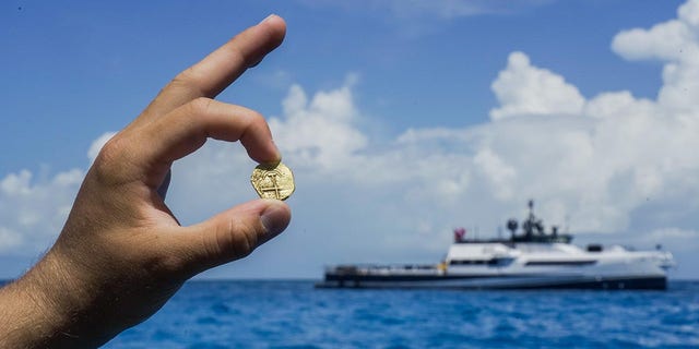 Исследователь держит золотую монету, найденную на Багамах, где исследовательское судно Аллена видно издалека.